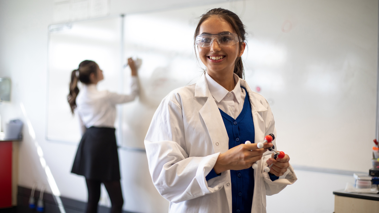 Two young people in a classroom. One is in the foreground holding a scientific model. The other writes on the whiteboard in the background.