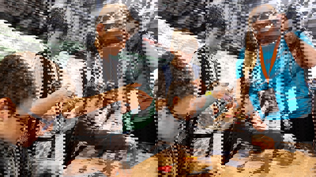 A selection of young people are working to complete a task with materials such as scissors, card, recyclable pots and paper clips. Some are sitting, some are standing. A volunteer offers hands on support. 