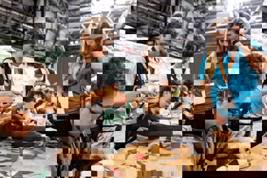 A selection of young people are working to complete a task with materials such as scissors, card, recyclable pots and paper clips. Some are sitting, some are standing. A volunteer offers hands on support.