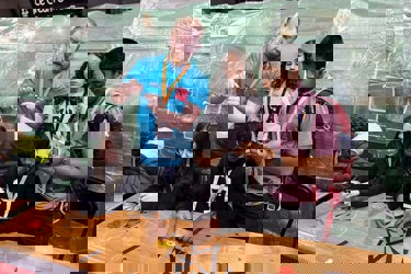 A volunteer at The Big Bang Fair demonstrates a task to four young people. Two of these people are sat at at table as they work on the task while the other two stand. All students are wearing lanyards.