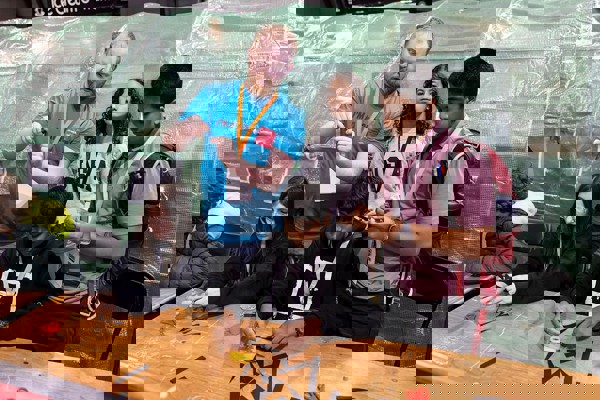 A volunteer at The Big Bang Fair demonstrates a task to four young people. Two of these people are sat at at table as they work on the task while the other two stand. All students are wearing lanyards.