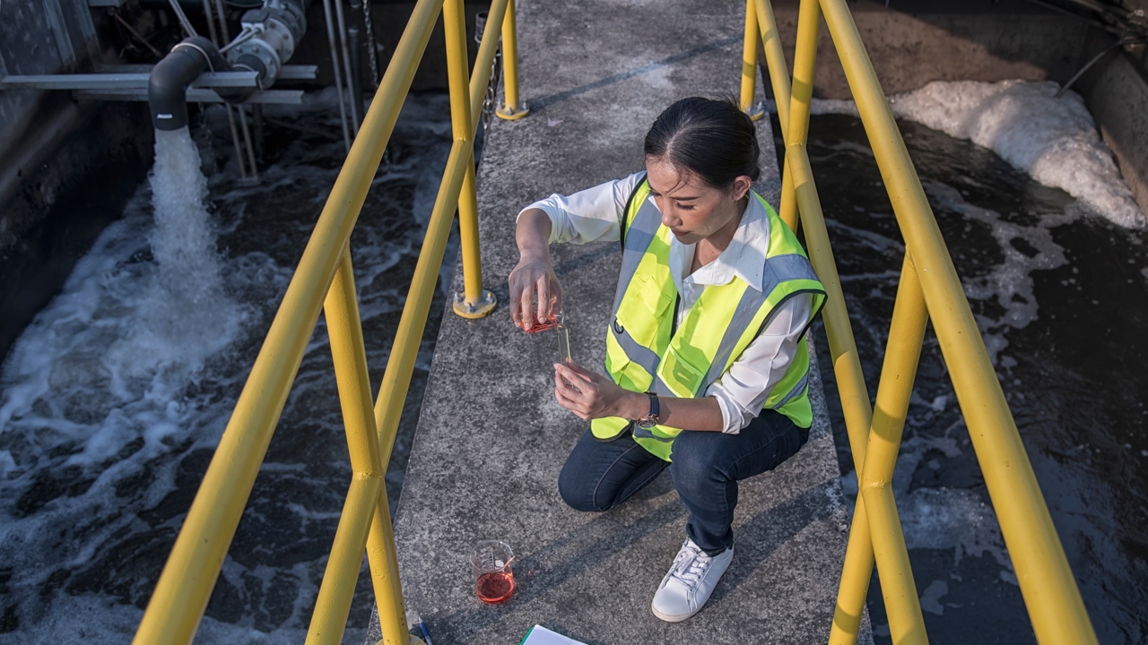 An engineer crouches on a footbridge over a body of water. They have beakers with chemicals. They wear high visibility outerwear. 