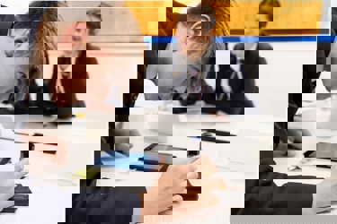 A school classroom. The focus is on one young person who is looking intently at a microscope as other members of the class appear in the background.