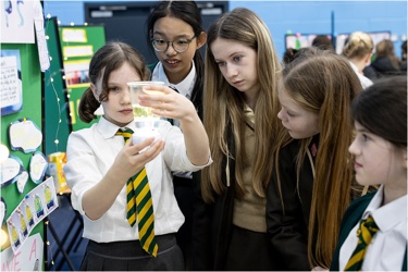 Five young people at a science fair, investigating a beaker of liquid. One holds the beaker as the others look on with interest.