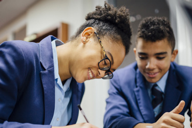 Secondary school students in a classroom at a desk working. One writes as the others look on.