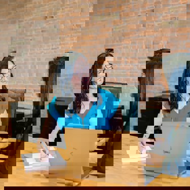 Two people sit at a large desk at work. They face each other. One has a pen and notepad, one has a laptop.