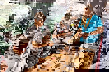 A selection of young people are working to complete a task with materials such as scissors, card, recyclable pots and paper clips. Some are sitting, some are standing. A volunteer watches on to support. 