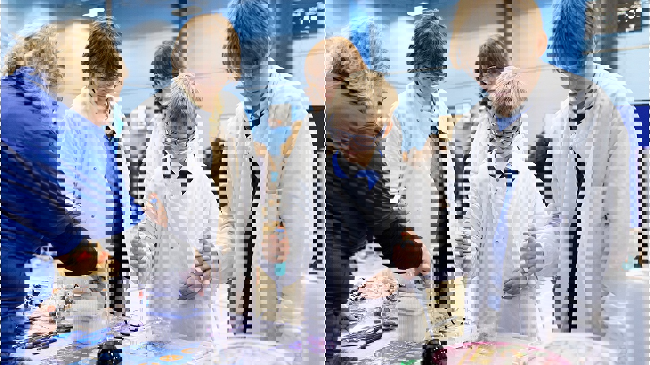 A school science fair setting. Four young people visit a stand and are conducting an experiment supported by a grown up. They all wear protective clothing and goggles. They work with electronic pipettes and liquids in beakers and flasks.