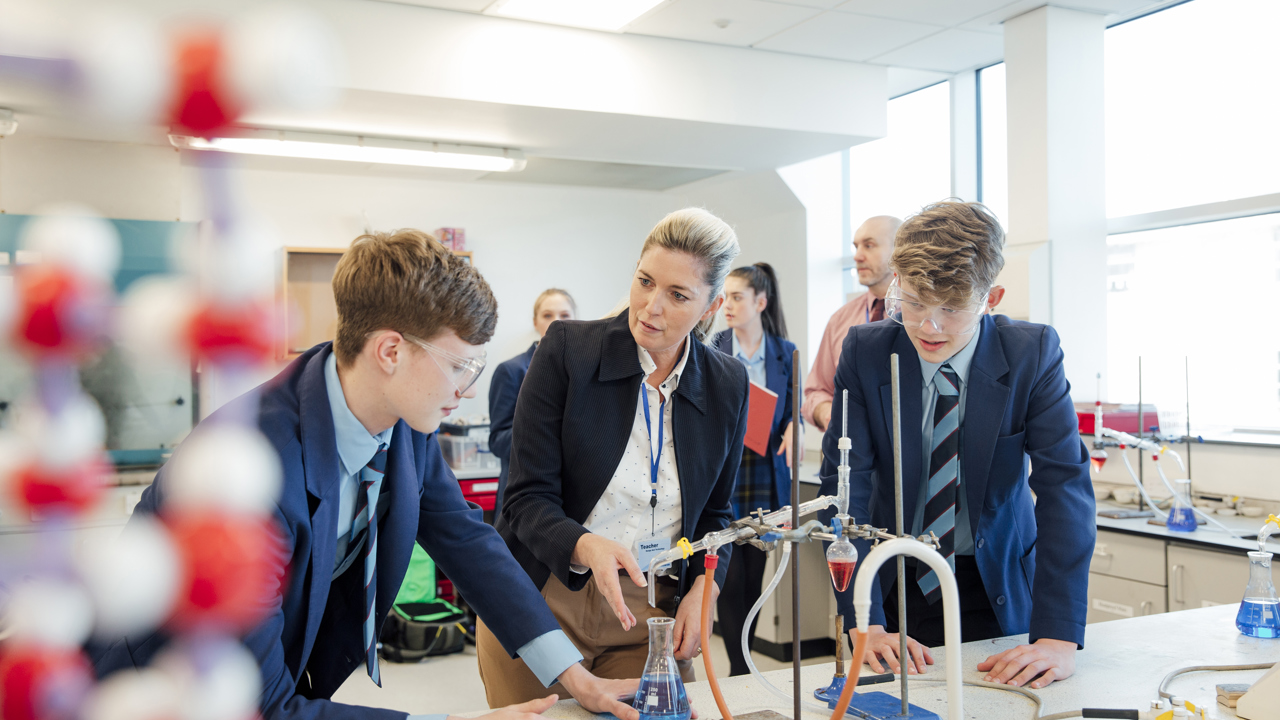 A school science lab where students are conducting an experiment with apparatus and protective equipment while a teacher looks on