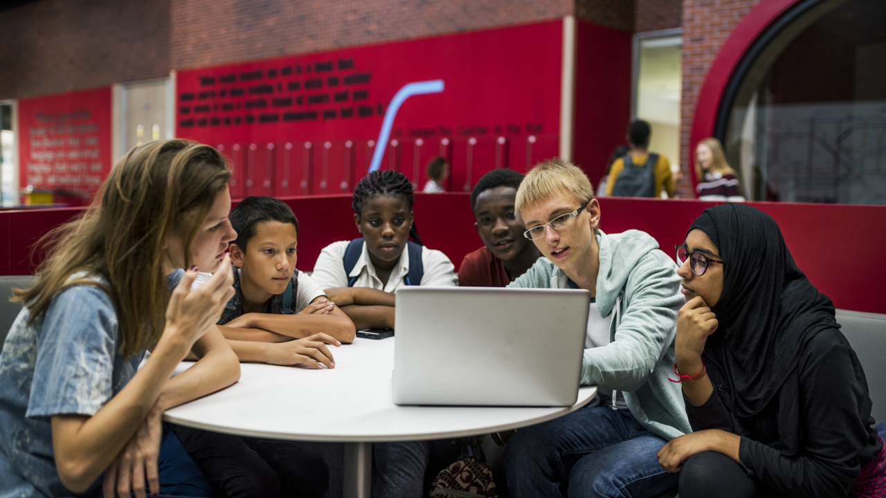 Six young people sit at a round table watching a laptop screen intently. 