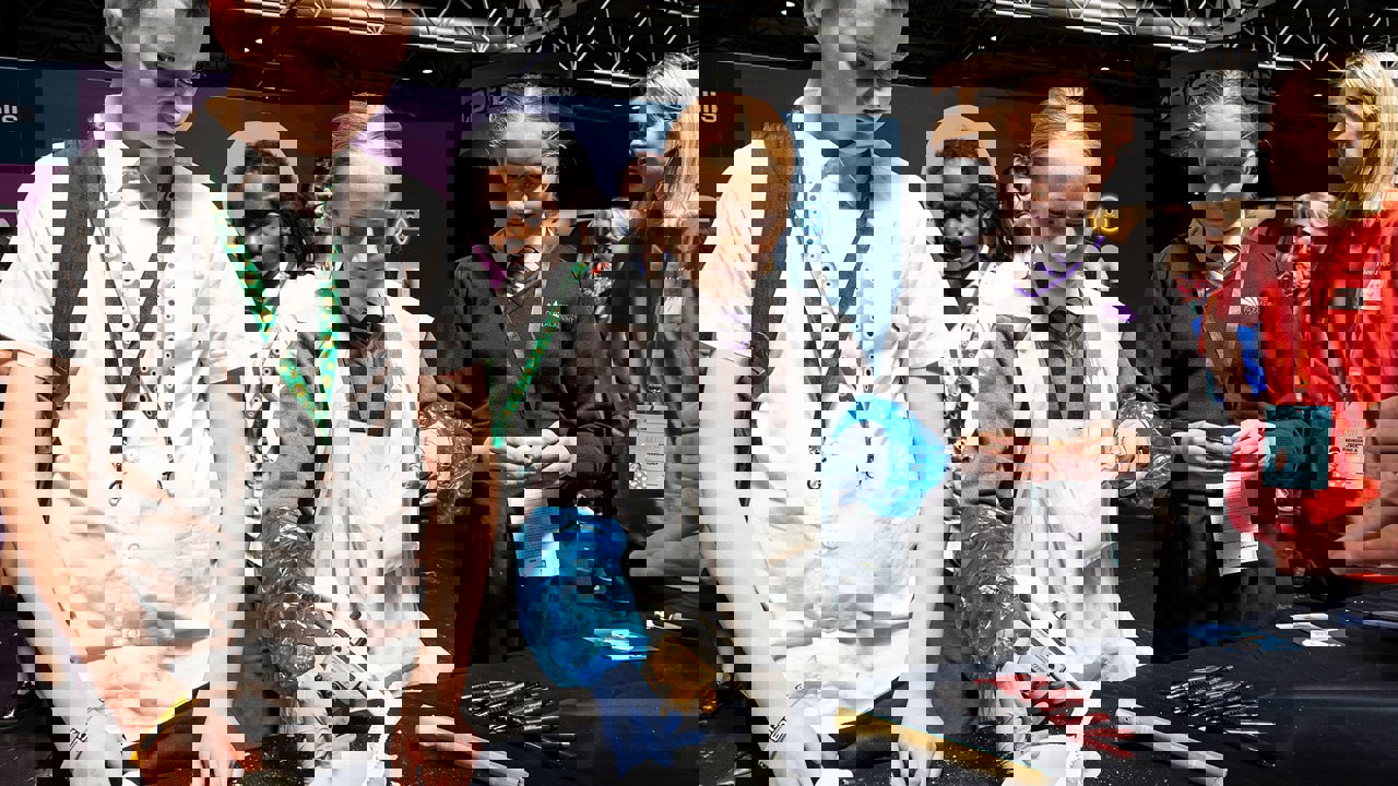 Several young people take part in a hands-on scientific experiment at The Big Bang Fair. One is wearing an apron and protective gloves as others watch. They are being supported by the adult on the stand.