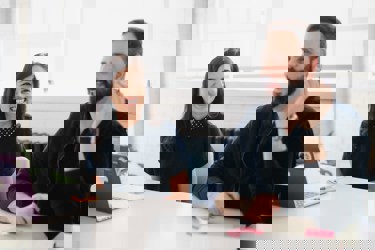 Two adults in an office meeting room setting. One has a laptop the other is talking. There are post-it notes on the desk alongside a laptop.