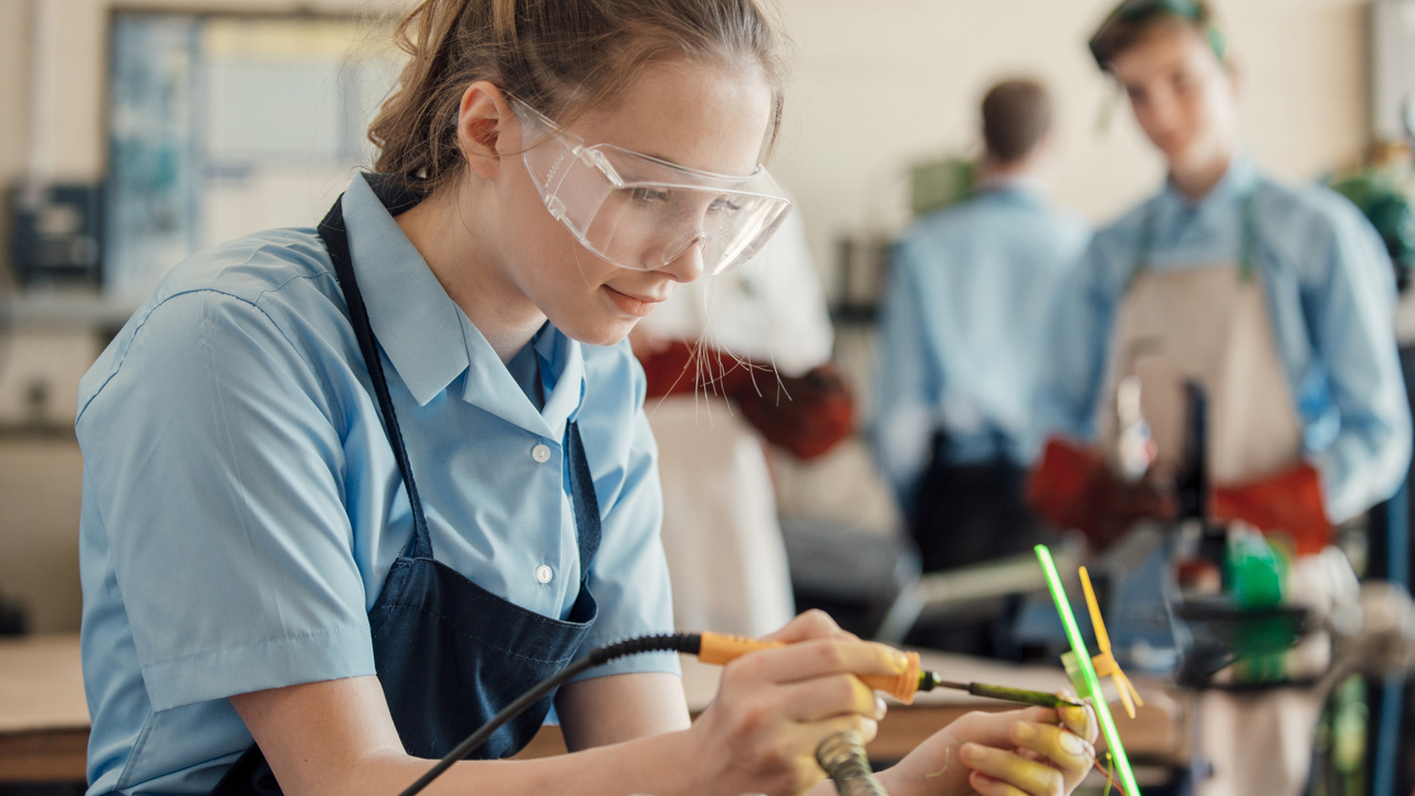 A secondary school student is in a classroom in a practical lesson. They are wearing protective goggles and apron as they work with electronic equipment, focusing on the materials on their workbench.