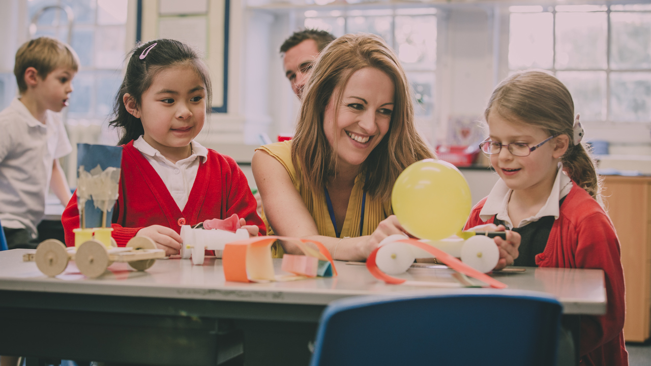 Primary school pupils in a classroom. A teacher sits with two pupils as they conduct a practical science experiment with wheels and balloons.