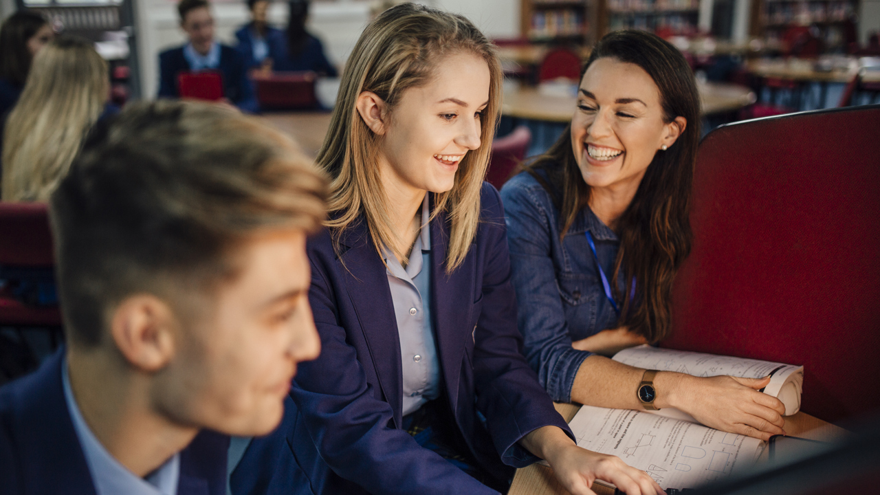 Secondary school students in a school library setting are in front of a screen being supported by their teacher. They have workbooks on the desk.