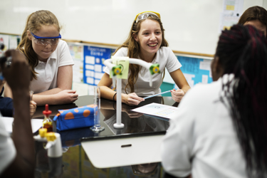 A group of secondary school students in a school science lab sit around a work bench with their pens and work book. There is scientific equipment on the surface too. The students are wearing protective goggles.
