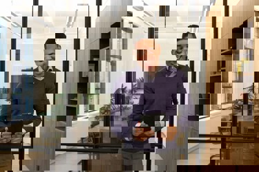 An adult stands in front of an empty boardroom with an iPad, looking animated