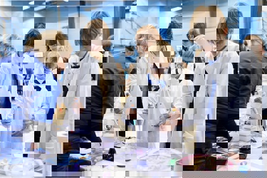 A school science fair setting. Four young people visit a stand and are conducting an experiment supported by a grown up. They all wear protective clothing and goggles. They work with electronic pipettes and liquids.