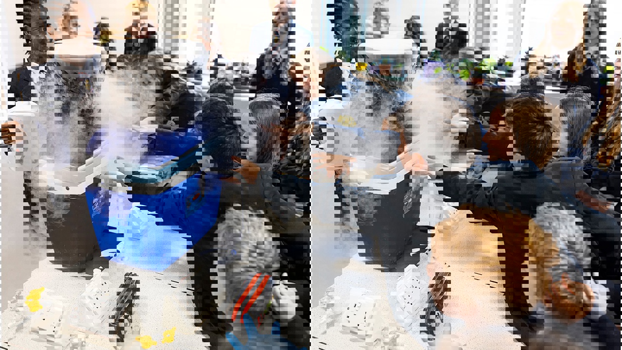 A school science lab. Many students surround a work bench as a cool box exudes dry ice. The students reach to touch it. They smile as they watch on. 