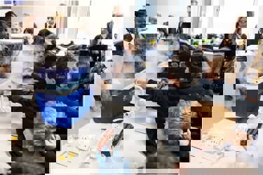 A school science lab. Many students surround a work bench as a cool box exudes dry ice. The students reach to touch it. They smile as they watch on.