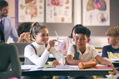 Primary school pupils in a classroom setting. The pupils have small model wind turbines on their desks along with text books and pencils. Two students are interacting with their model.