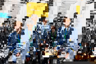 The six winners of The Big Bang Competition UK Young Technologists of the Year stand in an exhibition area displaying a model of their product along with sketches and working drawings to show how their idea developed. They all face the camera