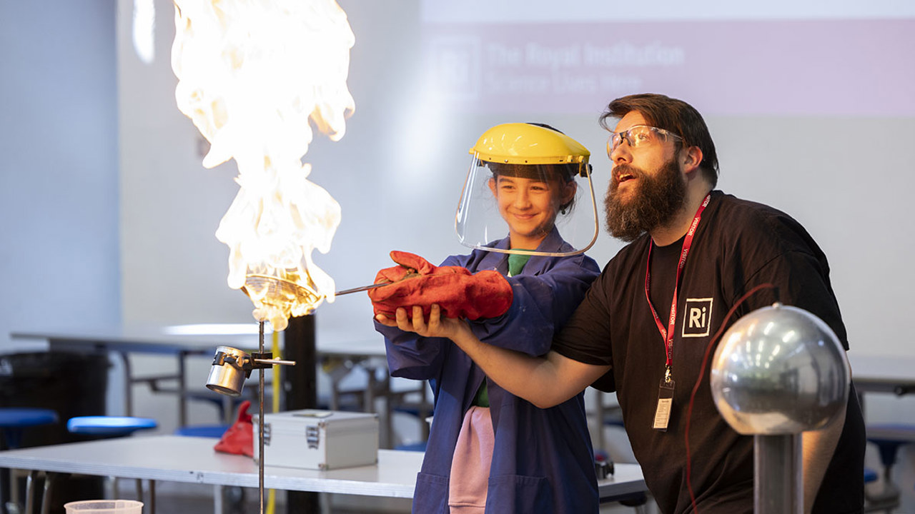 A young person and a teacher stand together conducting a scientific experiment with fire. The student has protective gloves, goggles and overall. They are holding something in the flame as the grown up supports them.