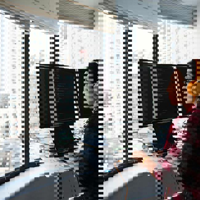 An adult sits at a desk with headphones on. They are typing on their laptop and looking at three screens. They are high up and outside their window you can see other buildings 