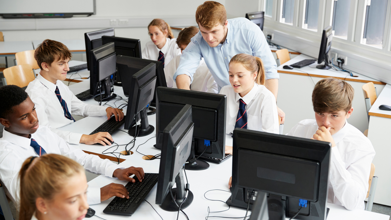 Seven students and a teacher work in a school computer lab. All the young people are looking at their individual screens on their desks.