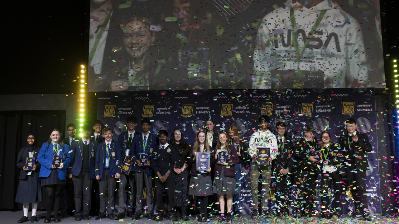 A group shot of The Big Bang Competition winners all together on a stage holding their awards. A confetti canon has released ticker tape which is still floating in the air, and some has rested on the stage.