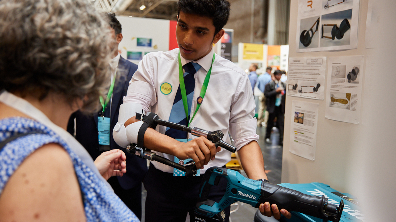A Big Bang Competition entrant shows their entry to a visitor to their stand at The Big Bang Fair. They are holding an implement to demonstrate how their idea works.